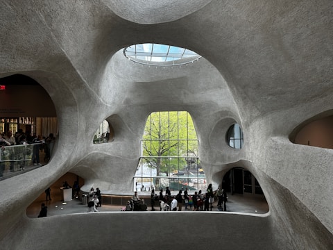 Futuristic architectural interior with organic, curving walls made of textured concrete. The space features large, irregular windows offering views of greenery outside. A glass ceiling allows natural light to illuminate the large open area, where groups of people are gathered, some looking out the windows and others congregating near an information desk.