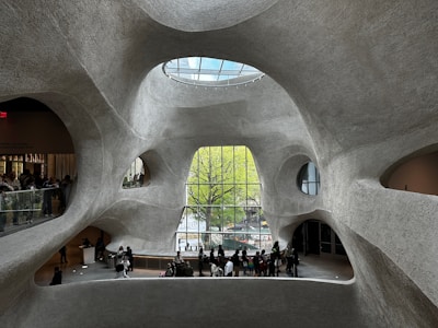 Futuristic architectural interior with organic, curving walls made of textured concrete. The space features large, irregular windows offering views of greenery outside. A glass ceiling allows natural light to illuminate the large open area, where groups of people are gathered, some looking out the windows and others congregating near an information desk.