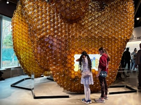 A large, translucent honeycomb structure dominates the scene, with two children interacting with a digital screen integrated into it. The honeycomb is composed of numerous hexagonal cells and is surrounded by a modern indoor environment.