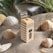 A biodegradable dental floss package made from corn fiber is placed on sand, surrounded by seashells and small plants. The packaging has a modern design with a houndstooth pattern and French text.