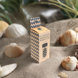 A biodegradable dental floss package made from corn fiber is placed on sand, surrounded by seashells and small plants. The packaging has a modern design with a houndstooth pattern and French text.