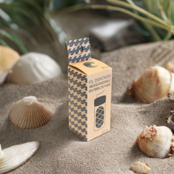 A biodegradable dental floss package made from corn fiber is placed on sand, surrounded by seashells and small plants. The packaging has a modern design with a houndstooth pattern and French text.