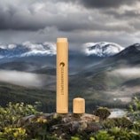 A bamboo tube labeled 'Oceans Respect' stands prominently on a rock, with a removed cap resting beside it. The background depicts a lush, expansive mountainous landscape under a cloudy sky, creating a striking contrast between the natural setting and the manufactured object.