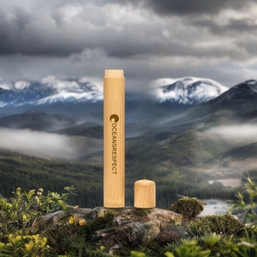A bamboo tube labeled 'Oceans Respect' stands prominently on a rock, with a removed cap resting beside it. The background depicts a lush, expansive mountainous landscape under a cloudy sky, creating a striking contrast between the natural setting and the manufactured object.