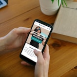 A person is holding a smartphone displaying a website with text promoting sustainable products and cleaner oceans. The background features a wooden table, a potted plant, and an open book.