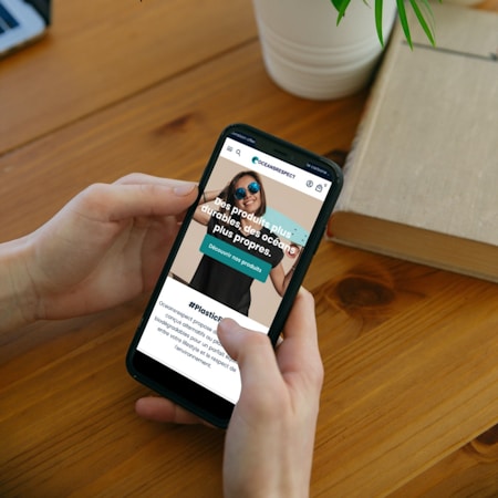 A person is holding a smartphone displaying a website with text promoting sustainable products and cleaner oceans. The background features a wooden table, a potted plant, and an open book.