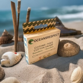 A box of bamboo cotton swabs labeled 'OCEANSRESPECT' on a sandy beach. The box is surrounded by seashells, and the ocean is visible in the background with gentle waves.