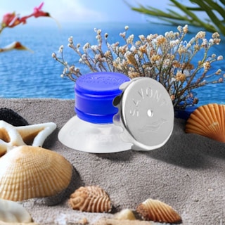 A coastal-themed arrangement with various seashells placed on sand. In the foreground is a blue suction cup device with a silver cap labeled 'Savont'. Behind the main objects, sprigs of dried flowers and a glimpse of the ocean with clear blue water are visible.