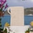 A woven beige pouch with a looped handle is positioned centrally against a backdrop of a serene blue sea and distant green hills. The scene is framed by vibrant flowers and lush greenery, with a white pedestal underneath the pouch.