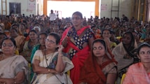 A large group of women seated in a hall, with one woman standing and speaking into a microphone. The women are dressed in colorful traditional attire, and appear attentive and focused on the person speaking.