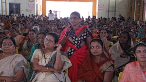 A large group of women seated in a hall, with one woman standing and speaking into a microphone. The women are dressed in colorful traditional attire, and appear attentive and focused on the person speaking.