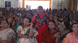 A large group of women seated in a hall, with one woman standing and speaking into a microphone. The women are dressed in colorful traditional attire, and appear attentive and focused on the person speaking.