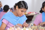 A person is carefully crafting or assembling pieces of jewelry, focusing intently on their work. The atmosphere is calm and appears to be in a workshop or classroom setting with others nearby.