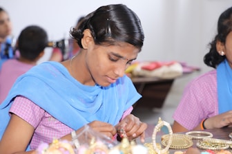 A person is carefully crafting or assembling pieces of jewelry, focusing intently on their work. The atmosphere is calm and appears to be in a workshop or classroom setting with others nearby.