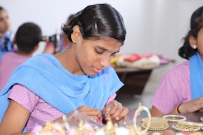 A person is carefully crafting or assembling pieces of jewelry, focusing intently on their work. The atmosphere is calm and appears to be in a workshop or classroom setting with others nearby.