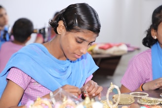A person is carefully crafting or assembling pieces of jewelry, focusing intently on their work. The atmosphere is calm and appears to be in a workshop or classroom setting with others nearby.