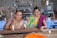 Two women are engaged in agricultural work, smiling as they handle soil or seeds on a metal sieve. They are wearing colorful sarees with traditional patterns, standing in a spacious indoor setting with machinery in the background.