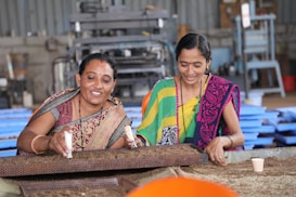 Two women are engaged in agricultural work, smiling as they handle soil or seeds on a metal sieve. They are wearing colorful sarees with traditional patterns, standing in a spacious indoor setting with machinery in the background.