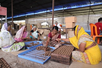 Several women dressed in colorful traditional clothing are sitting on the ground in a large indoor space, working with clay to create small handcrafted items. They are gathered around blue trays filled with clay pieces, interacting and focusing on their tasks. The background shows cardboard boxes and other people engaged in similar activities.