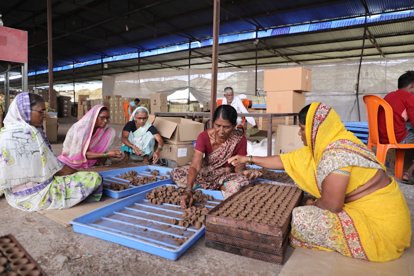 Several women dressed in colorful traditional clothing are sitting on the ground in a large indoor space, working with clay to create small handcrafted items. They are gathered around blue trays filled with clay pieces, interacting and focusing on their tasks. The background shows cardboard boxes and other people engaged in similar activities.