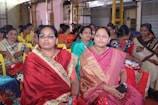 A group of women wearing colorful traditional saris are seated together in what appears to be a community or social gathering. They are smiling and engaged, with some holding items such as a phone and a small box. The setting is indoors, with metal shutters and a structural framework visible in the background.