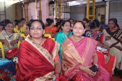 A group of women wearing colorful traditional saris are seated together in what appears to be a community or social gathering. They are smiling and engaged, with some holding items such as a phone and a small box. The setting is indoors, with metal shutters and a structural framework visible in the background.