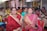 A group of women wearing colorful traditional saris are seated together in what appears to be a community or social gathering. They are smiling and engaged, with some holding items such as a phone and a small box. The setting is indoors, with metal shutters and a structural framework visible in the background.