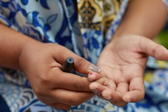 A close-up of hands, one holding a small black device while the other shows a fingertip with a small dot of blood. The background features a colorful patterned fabric.