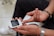A person is testing their blood sugar using a glucometer. The person's hand holds the device with a test strip inserted and a drop of blood on their fingertip. They are wearing a silver watch and a black wristband.