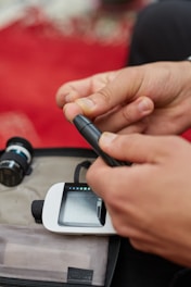 A person using a blood glucose meter, holding a lancet device to prick their finger. The kit includes a digital display screen and a test strip compartment. The background is a blurred red surface.