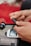 A person using a blood glucose meter, holding a lancet device to prick their finger. The kit includes a digital display screen and a test strip compartment. The background is a blurred red surface.