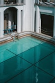 An empty swimming pool with clear turquoise water is surrounded by white walls and classic architectural elements, including columns and archways. A metallic blue railing provides access to the pool. The area is well-lit, suggesting daylight, and the shadows indicate the presence of overhanging structures.