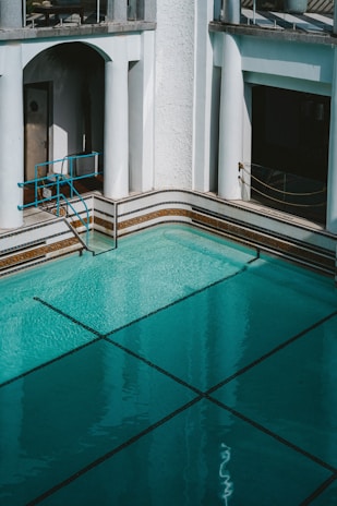An empty swimming pool with clear turquoise water is surrounded by white walls and classic architectural elements, including columns and archways. A metallic blue railing provides access to the pool. The area is well-lit, suggesting daylight, and the shadows indicate the presence of overhanging structures.