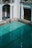 An empty swimming pool with clear turquoise water is surrounded by white walls and classic architectural elements, including columns and archways. A metallic blue railing provides access to the pool. The area is well-lit, suggesting daylight, and the shadows indicate the presence of overhanging structures.