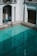 An empty swimming pool with clear turquoise water is surrounded by white walls and classic architectural elements, including columns and archways. A metallic blue railing provides access to the pool. The area is well-lit, suggesting daylight, and the shadows indicate the presence of overhanging structures.