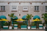 A building facade with a modern design, featuring large windows and a stone exterior. In front of the building are several green potted palm trees, under turquoise awnings, accompanied by yellow umbrellas, creating a vibrant street café setting.