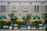 A building facade with a modern design, featuring large windows and a stone exterior. In front of the building are several green potted palm trees, under turquoise awnings, accompanied by yellow umbrellas, creating a vibrant street café setting.