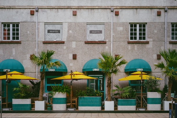 A building facade with a modern design, featuring large windows and a stone exterior. In front of the building are several green potted palm trees, under turquoise awnings, accompanied by yellow umbrellas, creating a vibrant street café setting.