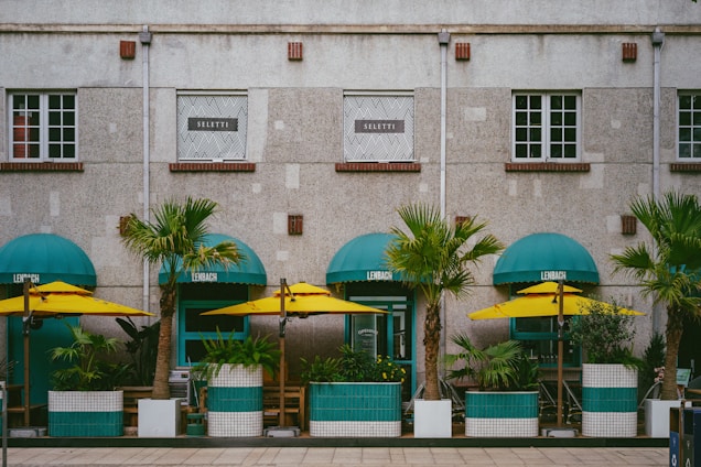 A building facade with a modern design, featuring large windows and a stone exterior. In front of the building are several green potted palm trees, under turquoise awnings, accompanied by yellow umbrellas, creating a vibrant street caf&eacute; setting.