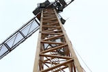 A towering construction crane is seen from a low angle, showcasing its lattice structure and industrial design against a pale sky. The crane appears to have rectangular lights and some cables running alongside it.