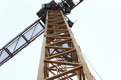 A towering construction crane is seen from a low angle, showcasing its lattice structure and industrial design against a pale sky. The crane appears to have rectangular lights and some cables running alongside it.