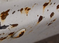 An aged, white-painted metal surface is heavily corroded, showing numerous rust spots and peeling areas. The rust has an irregular pattern and varies in intensity, ranging from dark brown to lighter orange shades. A round metal screw or button is visible, also affected by rust and corrosion.