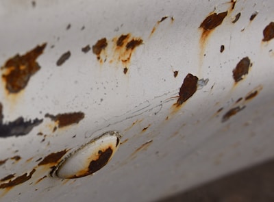 An aged, white-painted metal surface is heavily corroded, showing numerous rust spots and peeling areas. The rust has an irregular pattern and varies in intensity, ranging from dark brown to lighter orange shades. A round metal screw or button is visible, also affected by rust and corrosion.