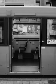 A bus is parked with its doors open, revealing a view of a few seats designated for disabled access inside. Outside, a group of workers in uniforms and helmets are visible near a truck, seemingly engaged in work or a discussion. The image is in black and white, emphasizing the tones and textures of the scene.