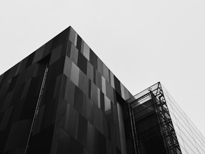 A modern architectural building with sharp, angular lines and a facade featuring a mix of solid and glass panels. The stark contrast between the dark building and the pale sky enhances its contemporary design.