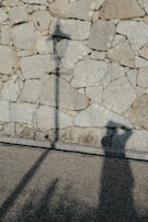 Shadows of a person with an outstretched arm, possibly holding something, are cast on a stone wall composed of uneven, rough-textured rocks. The shadows convey a sense of mystery and intrigue.