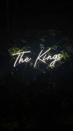 Neon sign spelling out 'The Kings' illuminated against a dark background with green leaves surrounding it.