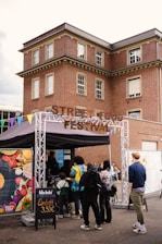 People are gathered in front of a booth with a sign reading 'Street Food Festival.' The booth has colorful triangular flags attached, and a variety of food can be seen in a poster on its side. A brick building with multiple windows serves as the backdrop.