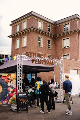 People are gathered in front of a booth with a sign reading 'Street Food Festival.' The booth has colorful triangular flags attached, and a variety of food can be seen in a poster on its side. A brick building with multiple windows serves as the backdrop.