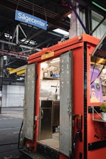A red food truck with open doors reveals a stainless steel interior, including a sink and shelving. Above, a blue sign reads 'Sägecenter' inside a large industrial warehouse with visible steel beams and pipes. On the right side of the truck, a poster with vivid graphics adds a pop of color.